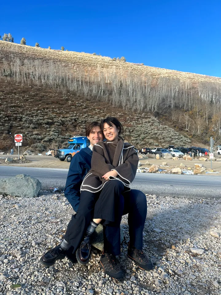 Logan and his partner sitting on a rock at the top of Guardsman Pass on a sunny fall day, with aspens and sagebrush on the hillside behind them.